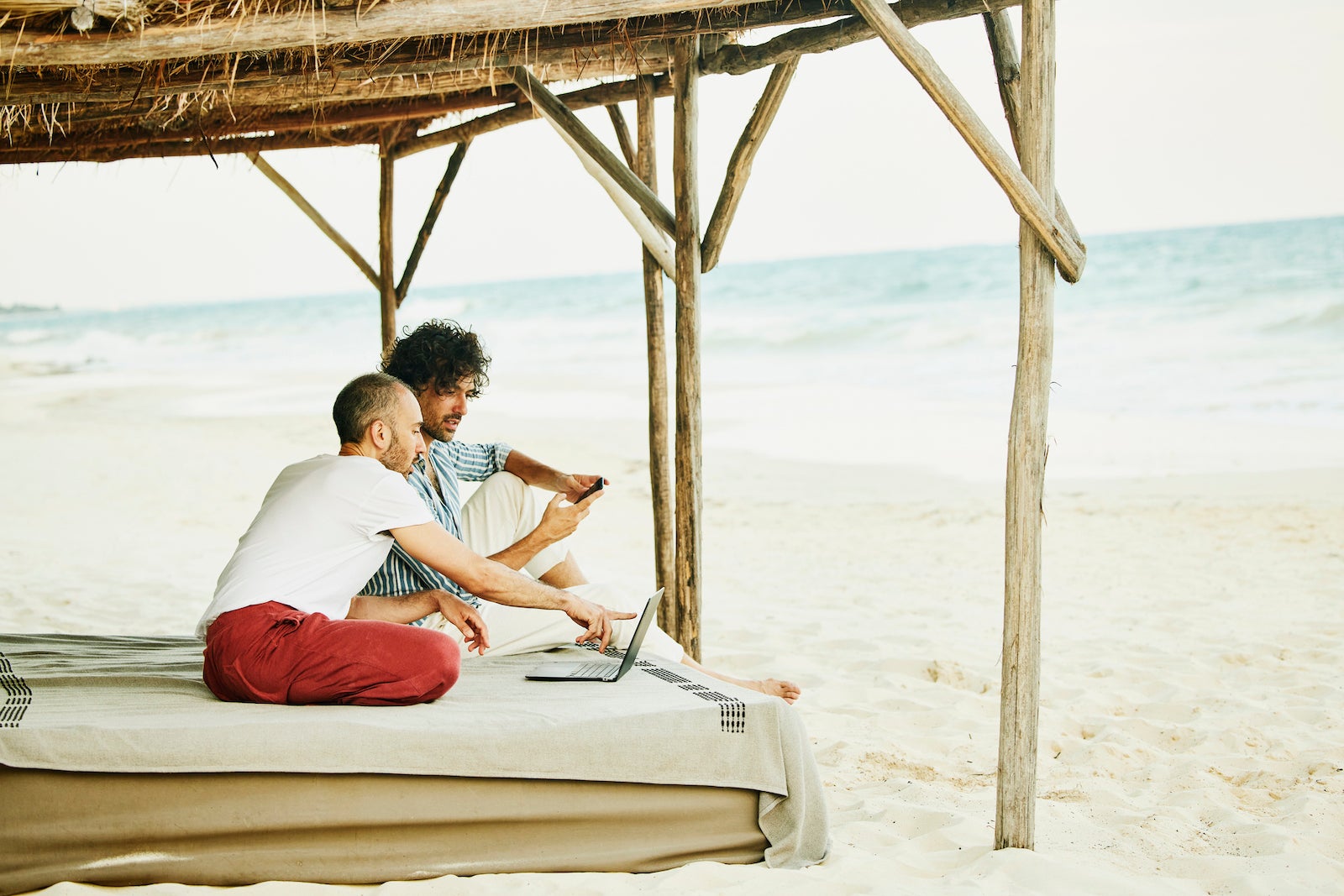 Wide shot of gay couple looking at data laptop while relaxing at beach cabana at tropical resort